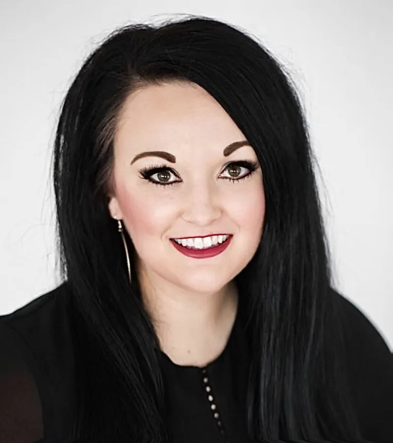 Smiling woman with long black hair, bold brows, and dark red lips against a light gray background