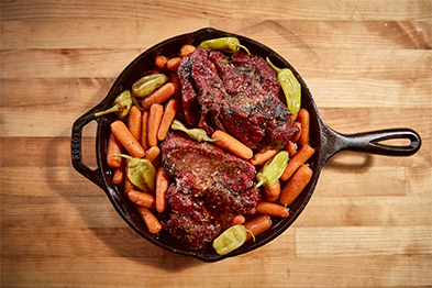 Cast iron skillet with braised meat, baby carrots, and peppadew peppers on a wood surface