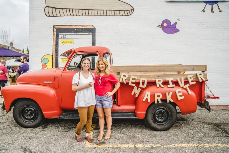 Two women posing in front of a vintage red truck with a Red River Market banner