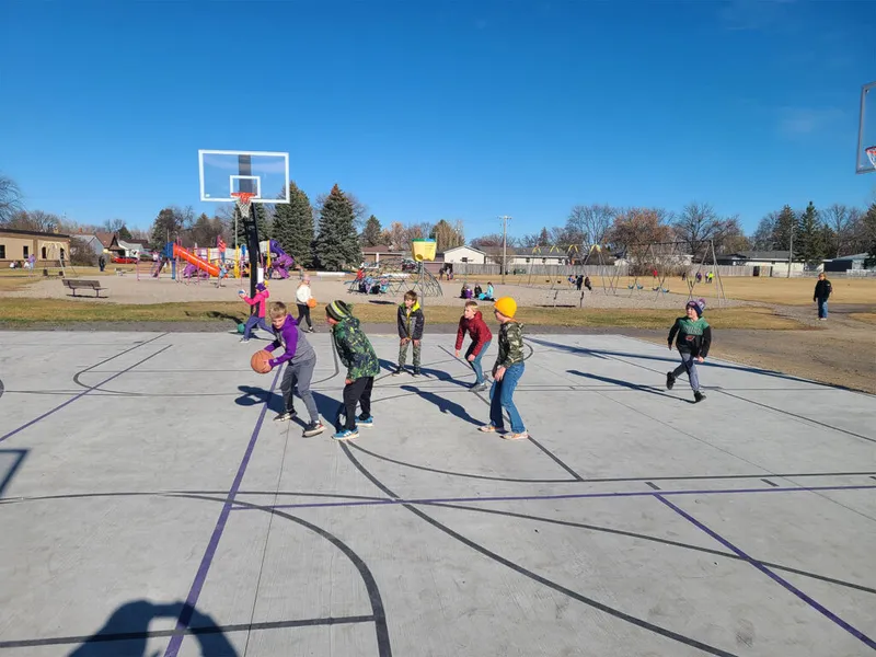 Children in winter jackets playing basketball on an outdoor court next to a school playground