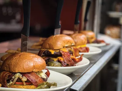 Row of loaded bacon cheeseburgers on white plates with fries in background