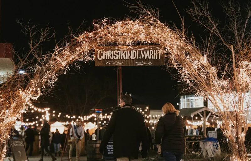 Lit twig arch with Christkindlmarkt sign over crowd at a night market