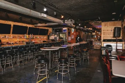 Dimly lit bar interior with metal barstools, high-top tables, and a fully stocked back bar