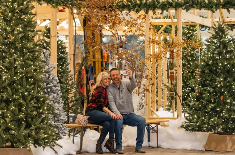 Couple taking a selfie on a bench surrounded by lit Christmas trees in a festive holiday display