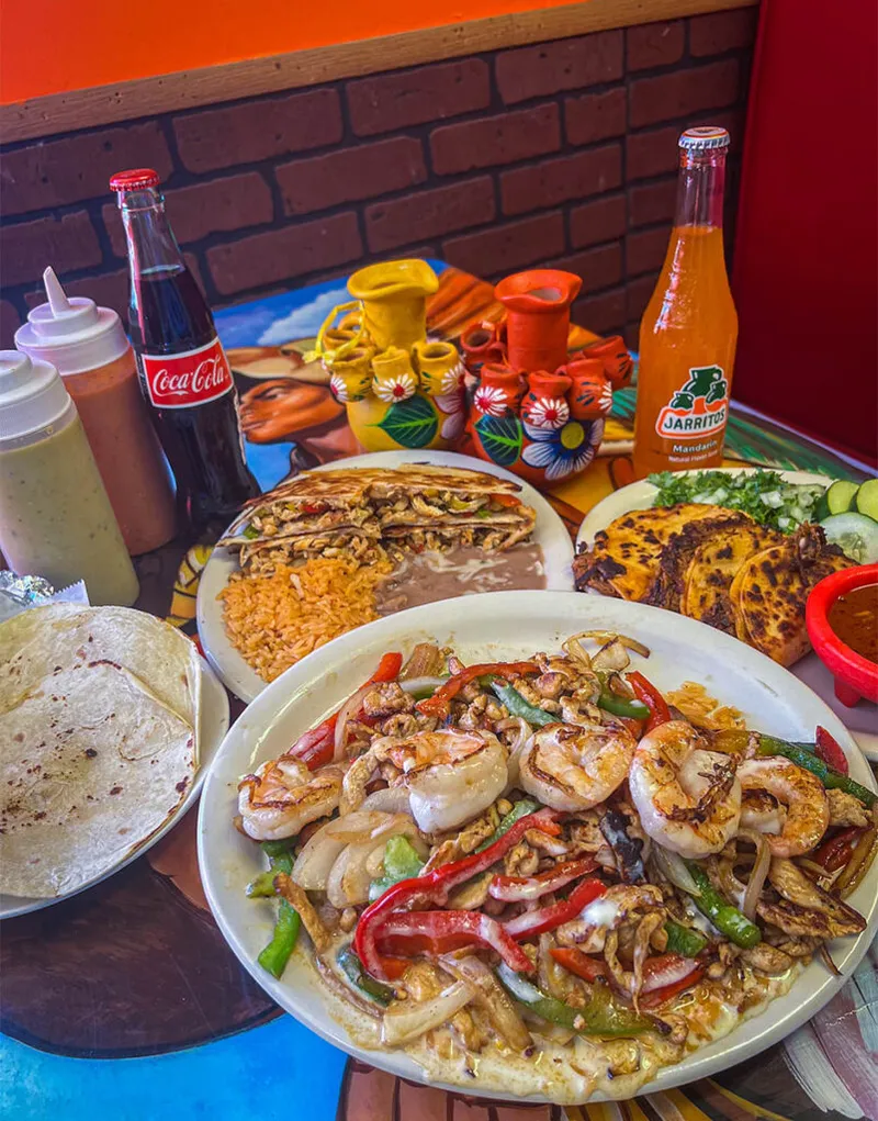 Shrimp fajitas with peppers and onions, a quesadilla plate with rice and beans, Coca-Cola, and Jarritos on a colorful table