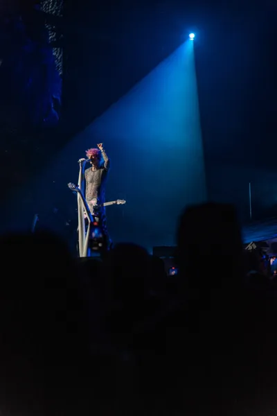 Pink-haired performer on stage beneath a single blue spotlight above a dark crowd