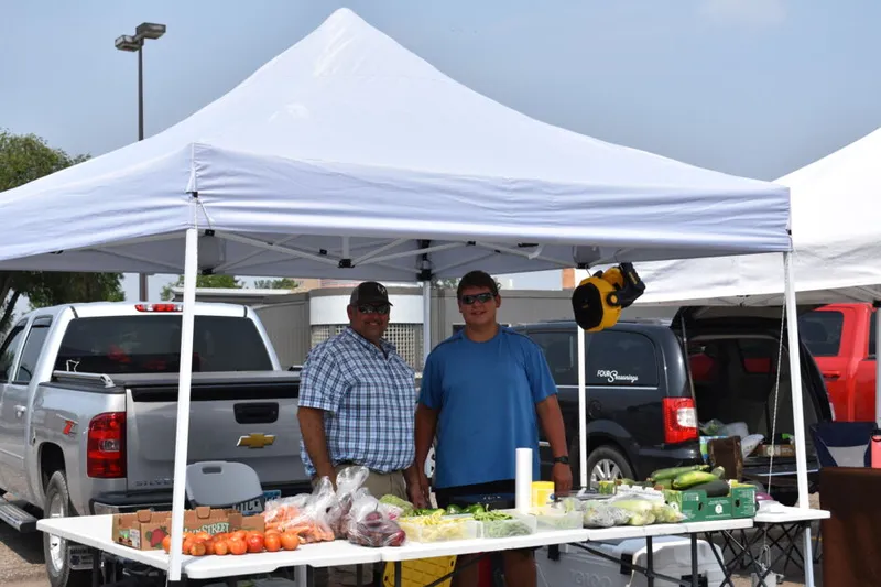 Two men standing behind a produce-laden table under a white canopy tent at an outdoor market