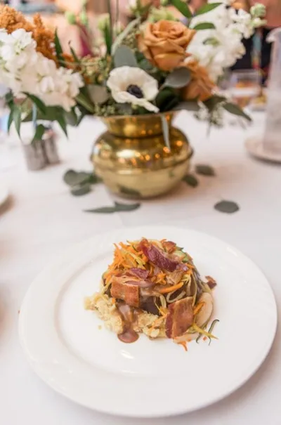 Plated dinner dish with sautéed vegetables and bacon on a white plate, gold vase with roses behind