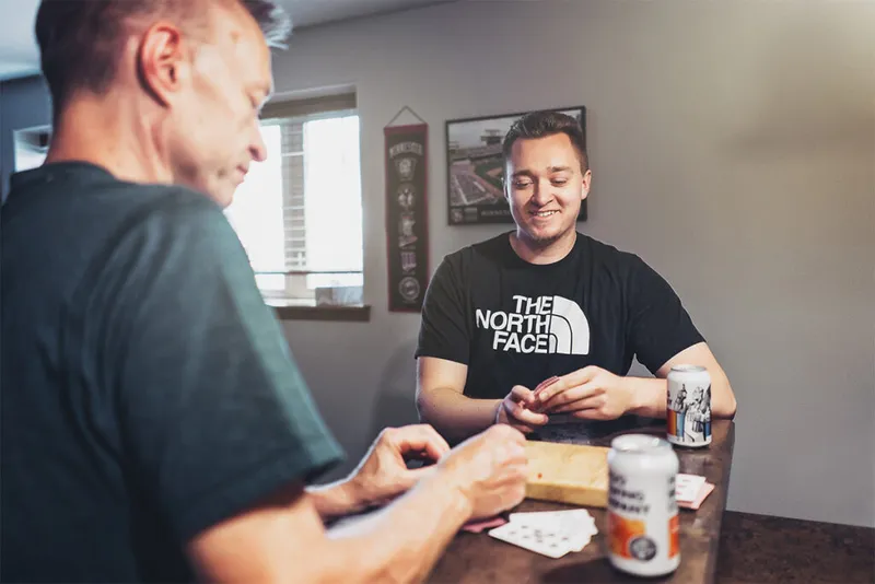 Two men playing cards at a table with beer cans nearby