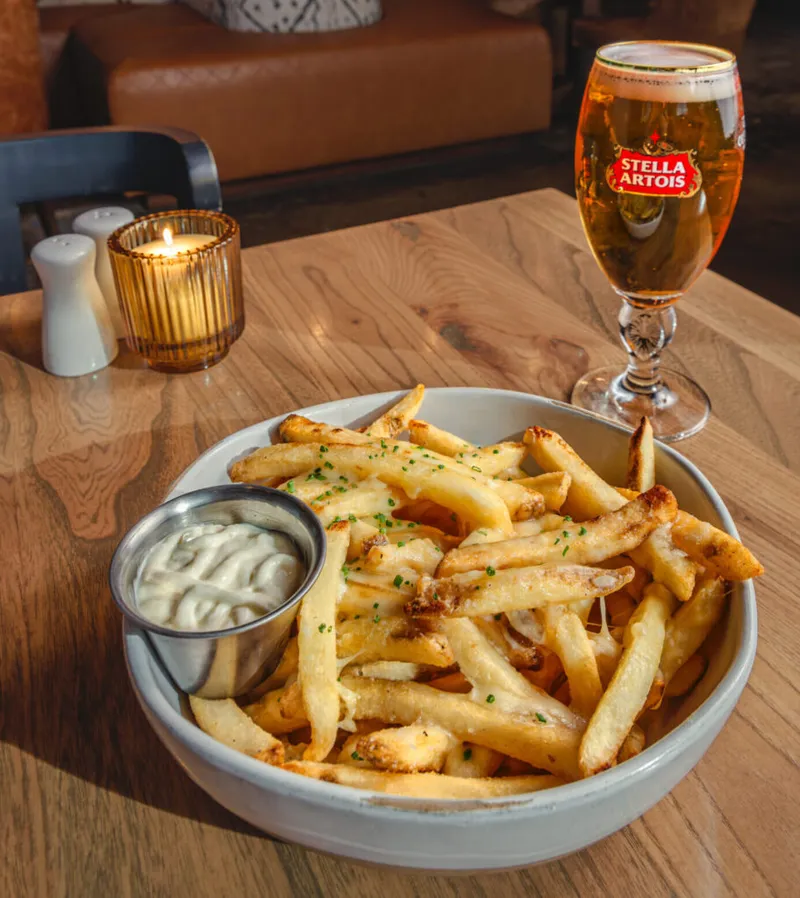 Cheesy fries with chive and dipping sauce beside a Stella Artois glass on a wood table