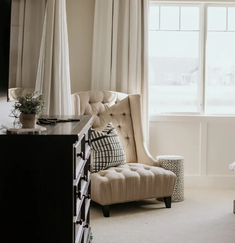 Cream tufted wingback chair with black and white check pillow beside a dark dresser and bright window