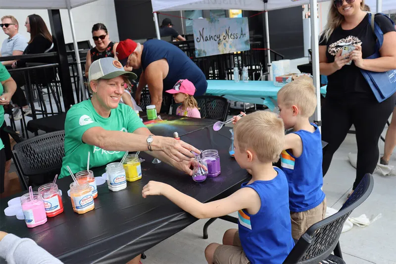 Woman in green shirt helps two young boys with colorful paint jars at an outdoor art activity table