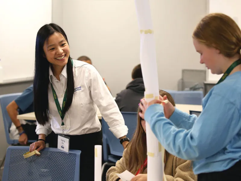 Smiling woman in white shirt and lanyard engages with students building a tall paper tube structure in a classroom