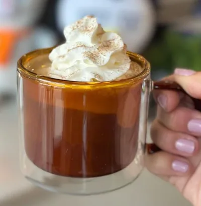 Hand holding a double-walled glass mug of dark coffee topped with whipped cream and cinnamon