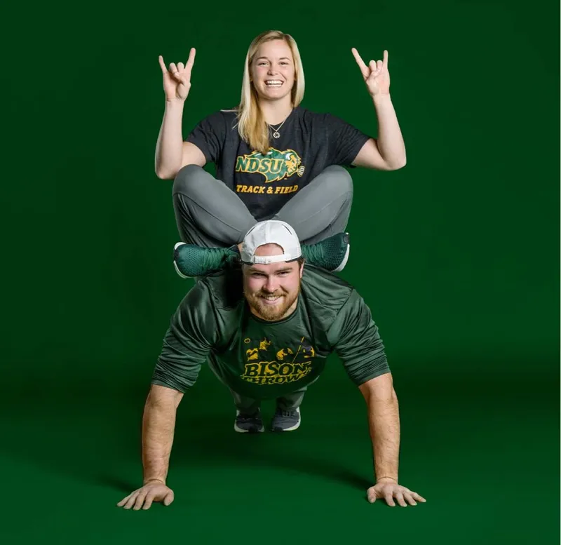 Two NDSU athletes in green and gold gear pose playfully against a green backdrop, one doing a headstand