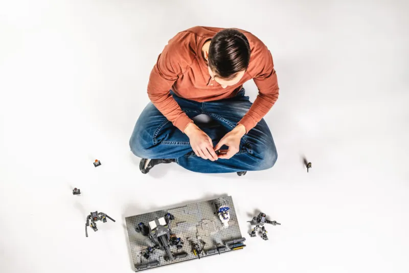 Man in rust-colored sweater sitting cross-legged on white floor, assembling dark-colored LEGO figures and a baseplate set