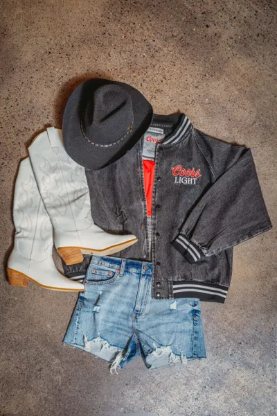 Flat lay of white cowboy boots, gray felt hat, distressed denim shorts, and a Coors Light varsity jacket