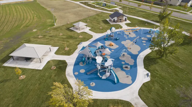 Aerial view of a modern playground with blue rubber surfacing and a nearby open-air pavilion on green grass