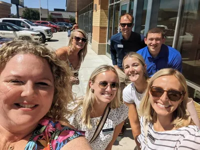Six smiling adults pose for an outdoor selfie near a strip mall parking lot
