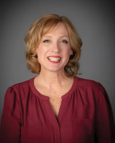 Smiling woman with short auburn hair wearing a dark red blouse against a gray background