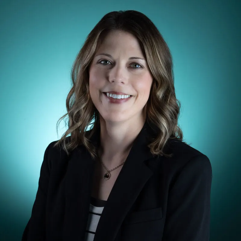 Smiling woman with wavy brown hair in a black blazer against a teal background
