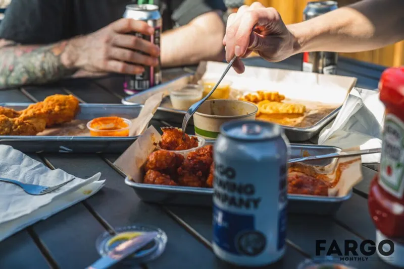 Hands reaching over an outdoor table spread with fried chicken, sauces, and canned drinks