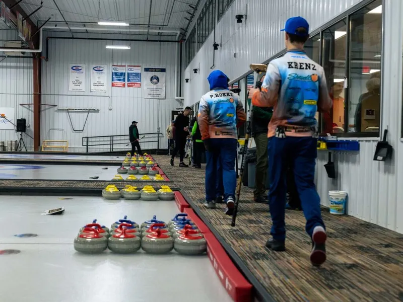 Curling players in matching jerseys walk past rows of red and yellow stones along an indoor ice sheet