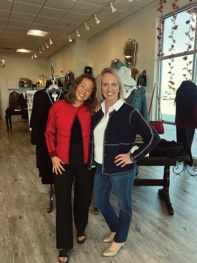 Two smiling women pose together inside a clothing boutique with racks of garments behind them