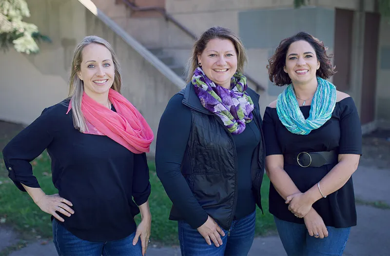 Three smiling women in black tops and colorful scarves posing outdoors