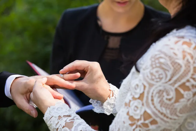 bride putting her groom's wedding ring on his finger during the ceremony with the officiant in the background