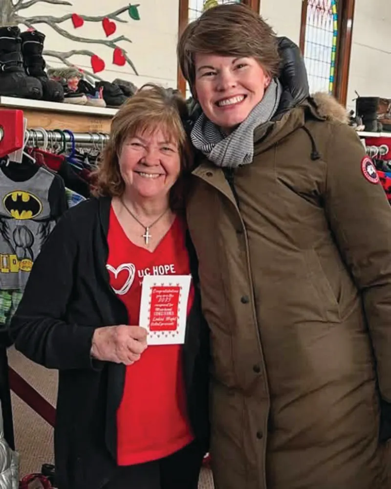 Two smiling women pose together inside a clothing donation space, one holding a congratulations card