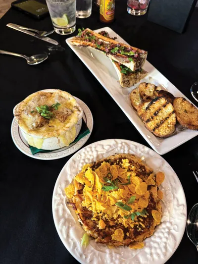 Three restaurant appetizers on a black tablecloth including a dip topped with crushed chips, a crock of French onion soup