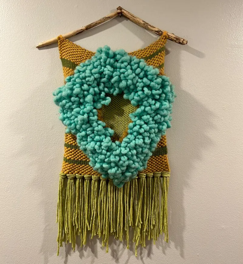 Woven wall hanging in mustard and teal with loopy yarn texture and green fringe on a driftwood rod