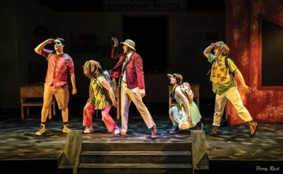 Six performers in colorful costumes strike dynamic poses on a theatre stage before an elementary school backdrop