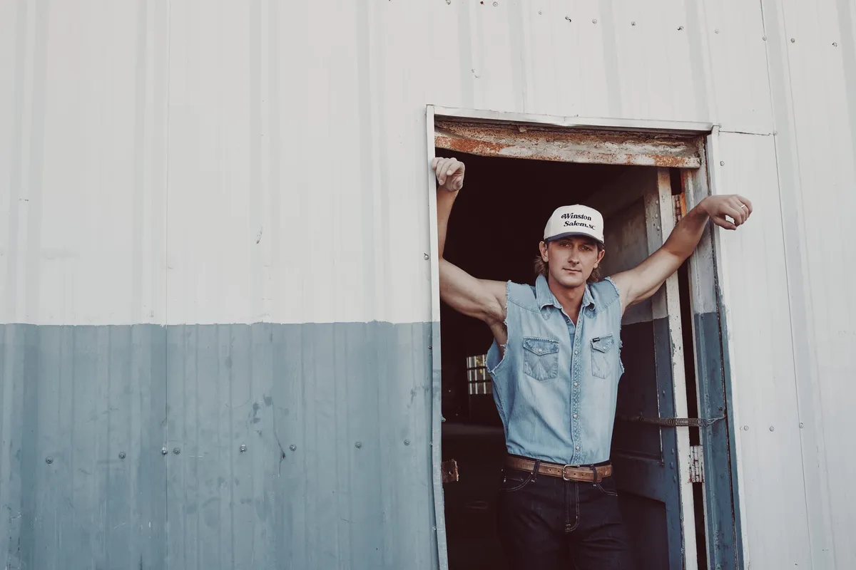 Man in sleeveless denim shirt and Winston-Salem trucker cap leaning in a metal barn doorway