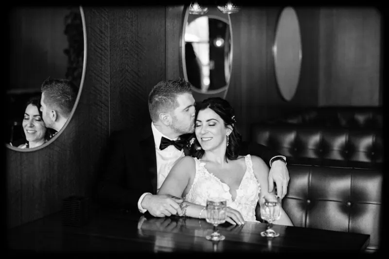 Groom in bow tie kisses bride's cheek in a dark leather booth, their reflection visible in a round mirror