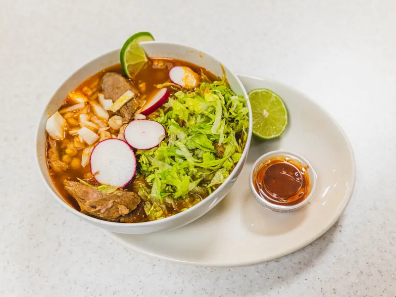 Bowl of red pozole topped with shredded cabbage, radish slices, diced onion, and lime, served with hot sauce