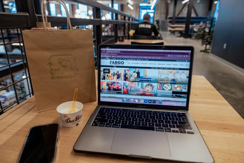 Laptop showing Fargo magazine website on a wood table beside a Duchess ice cream cup and kraft paper bag