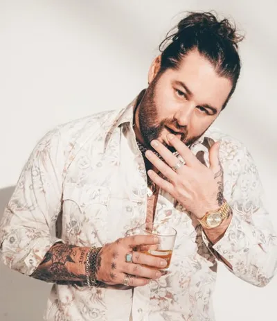 Tattooed bearded man with hair bun holding a glass, wearing a floral shirt and gold watch