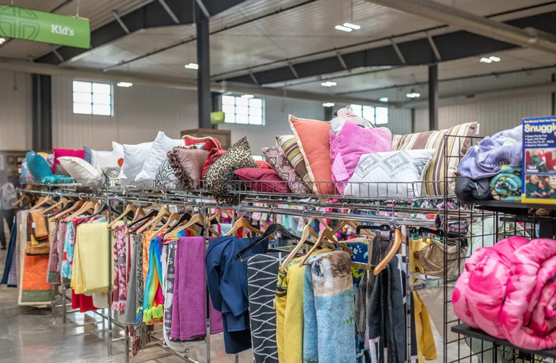 Colorful clothing on racks beneath shelves piled with decorative pillows in a thrift store