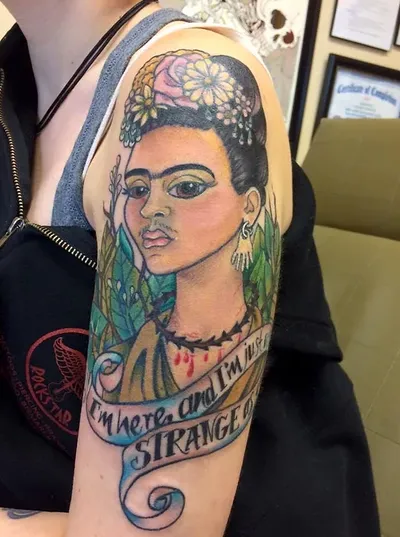 Colorful upper-arm tattoo of a woman with flowers in her hair and a banner reading 'I'm here and I'm strange