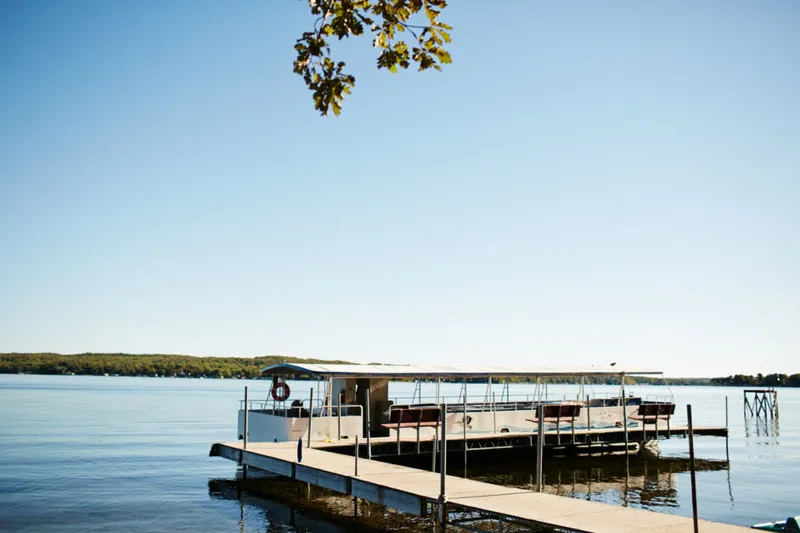 A covered pontoon boat docked at a wooden pier on a calm lake under clear blue skies