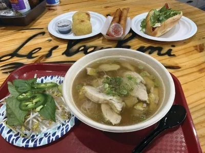 Bowl of chicken pho with fresh herbs beside egg rolls and a banh mi on a wooden table