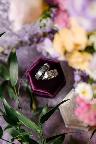 Two silver wedding bands in an open burgundy velvet hexagonal ring box surrounded by flowers
