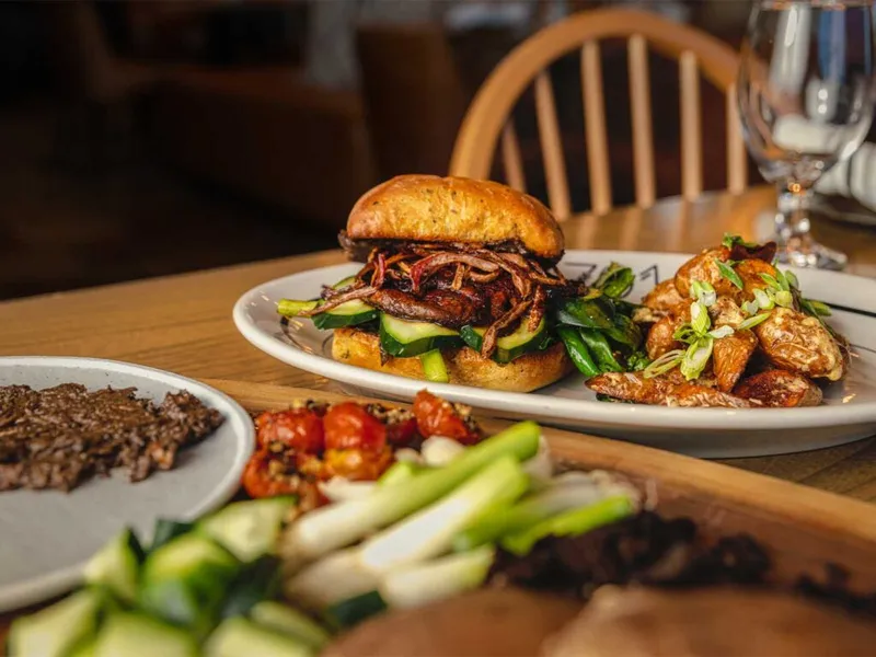 Pulled-meat sandwich with greens on a white plate, roasted potatoes alongside, and shared dishes on a wooden table