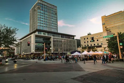 Busy downtown plaza at dusk with pedestrians, white event tents, and a tall modern glass building