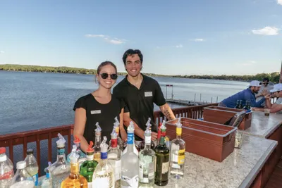 Two bartenders in black shirts smile behind an outdoor bar overlooking a lake