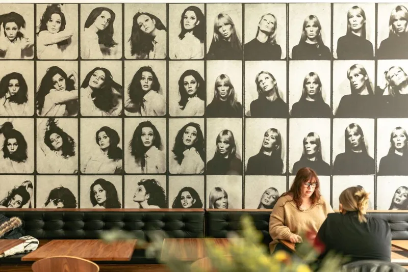 Two women chat in a restaurant booth beneath a wall mural of vintage black-and-white portrait photos