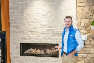 Smiling man in blue vest holding a remote beside a lit gas fireplace with stone surround