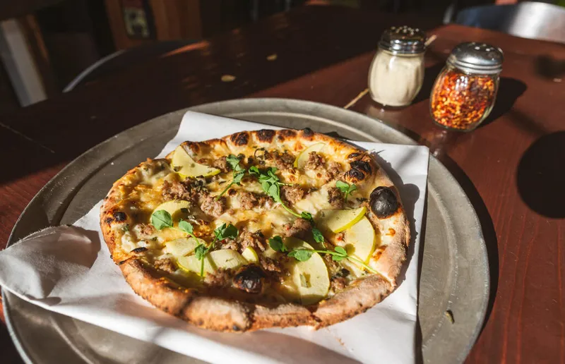 Wood-fired pizza topped with sliced pear, crumbled sausage, olives, and microgreens on a metal pan